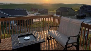 A deck with a grill and a chair, overlooking a rural landscape with houses and a sunset.