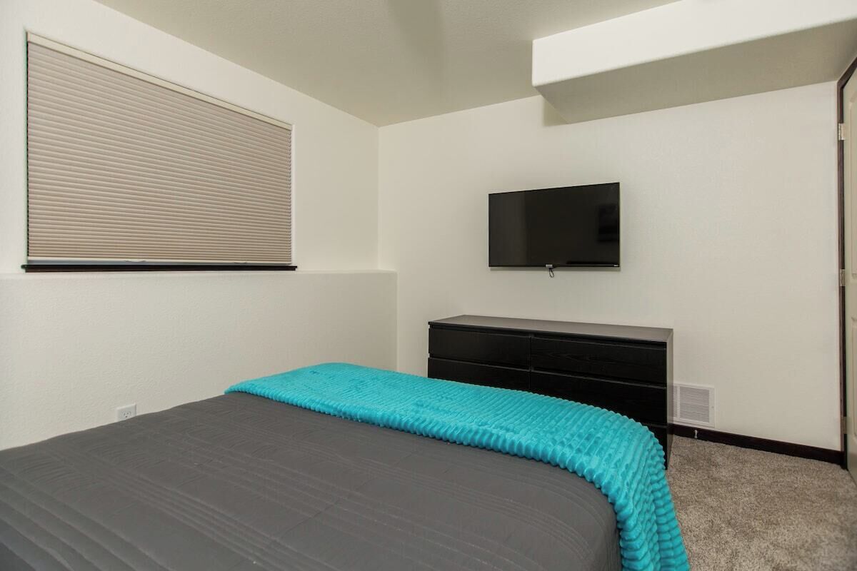 A bedroom with a bed, a TV mounted on the wall, and a window with blinds.