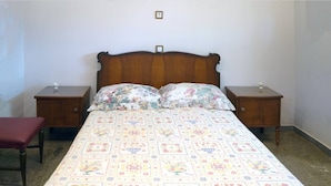 Un dormitorio con cabecera de madera, dos mesitas de noche, una colcha estampada con motivos florales y un banco rojo.