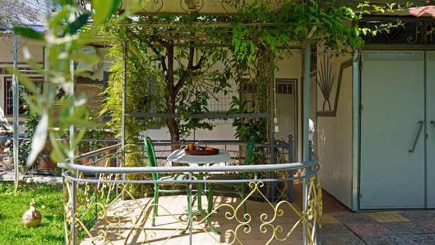 Un balcón con barandilla metálica, una mesa y sillas. Se ve un árbol con hojas verdes al fondo.