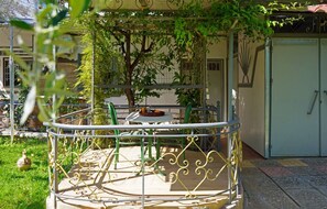 A balcony with a metal railing, a table, and chairs. There is a tree with green leaves in the background.