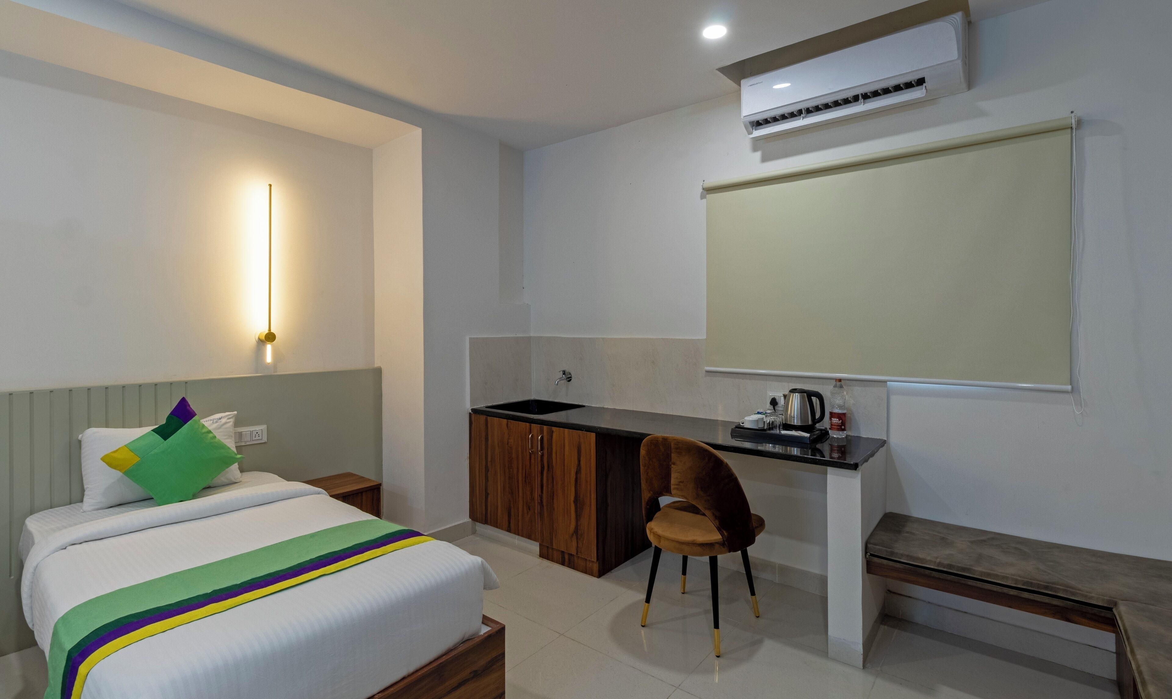 A hotel room with a bed, a desk, a chair, a sink, and an air conditioning unit.