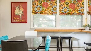A dining area with a glass table, black chairs, and a window with colorful floral blinds. - Eclectic Cottage by the bay, downtown, bike trail (Traverse City)