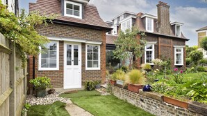 A brick house with a white door, a small garden with various plants, and a gravel path.