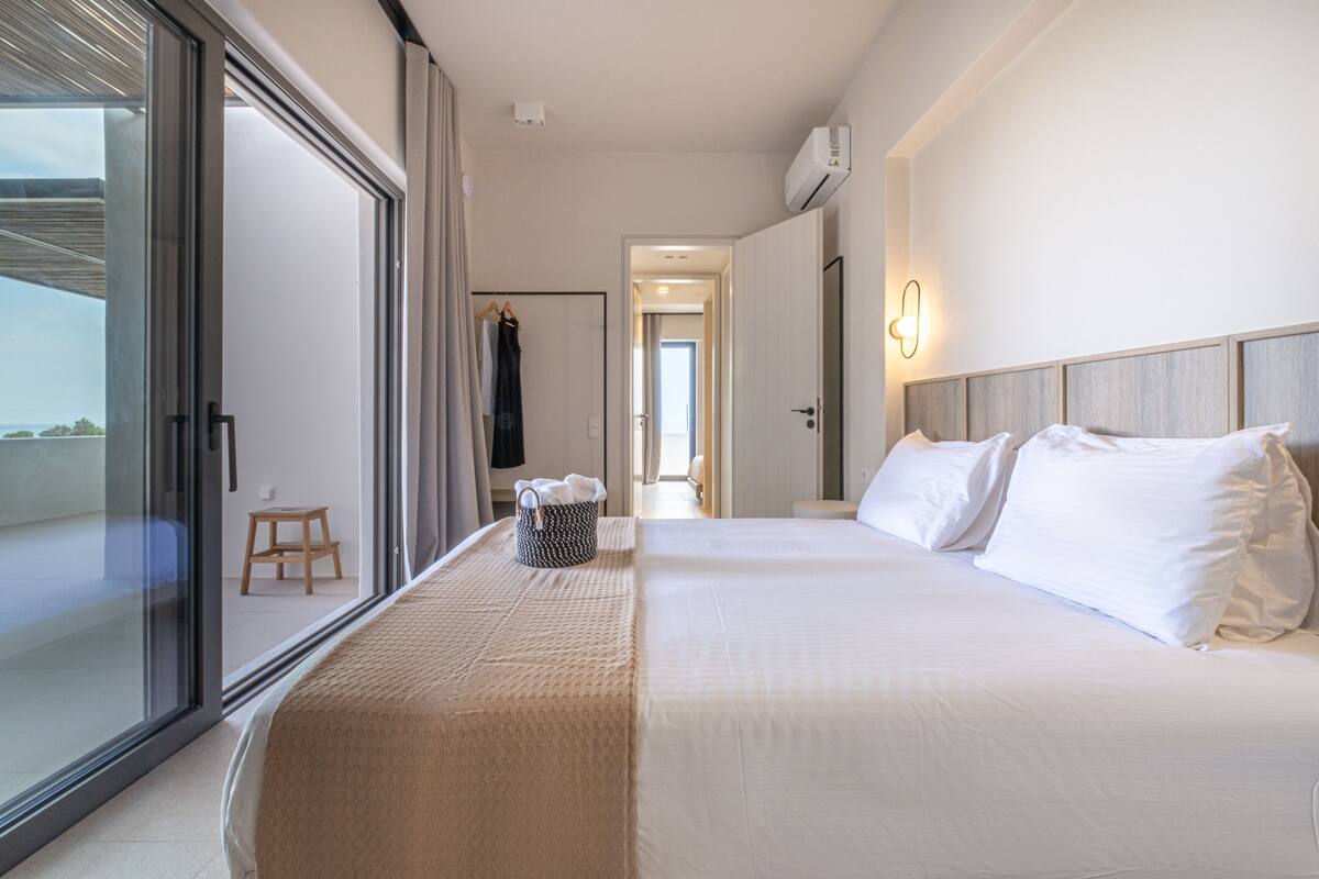 Een hotelkamer met een groot bed, een glazen schuifdeur naar een balkon en een klein houten krukje.