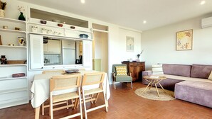 Ein modernes Wohnzimmer mit einer Couch, Sesseln, einem Esstisch und einer Küchenzeile mit Kühlschrank und Regalen.
