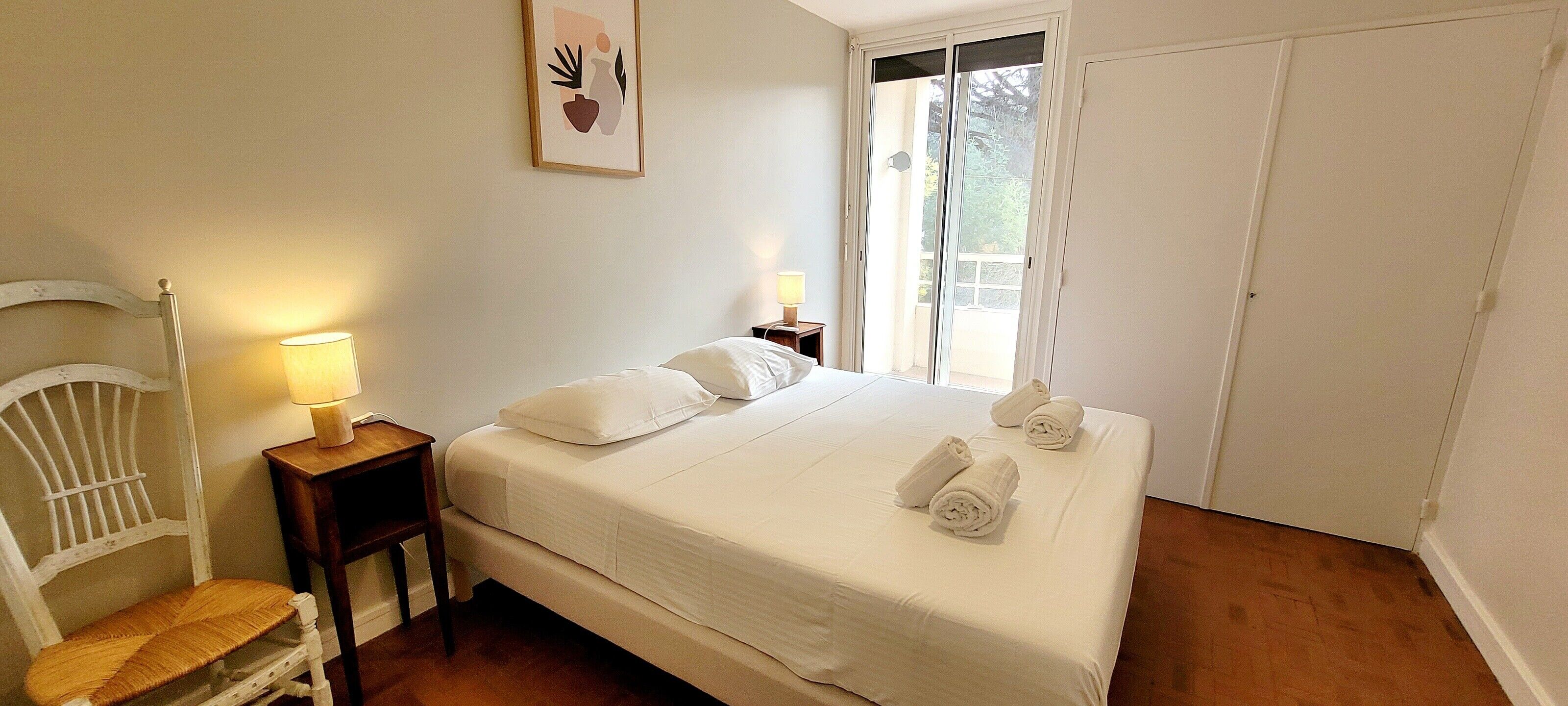 Ein Schlafzimmer mit einem Bett, einem Sessel, einem Nachttisch mit Lampe und einer Schiebetür zum Balkon.