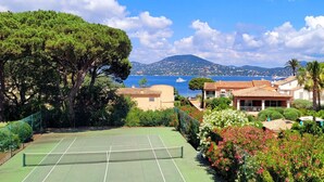 Ein Tennisplatz, umgeben von üppiger Vegetation und Gebäuden im mediterranen Stil, mit Blick auf das Meer und die Berge im Hintergrund.