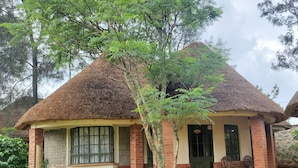 A small, single-story building with a thatched roof, brick walls, and a small porch with two chairs and a table. - Edaala Comfort (Nairobi)