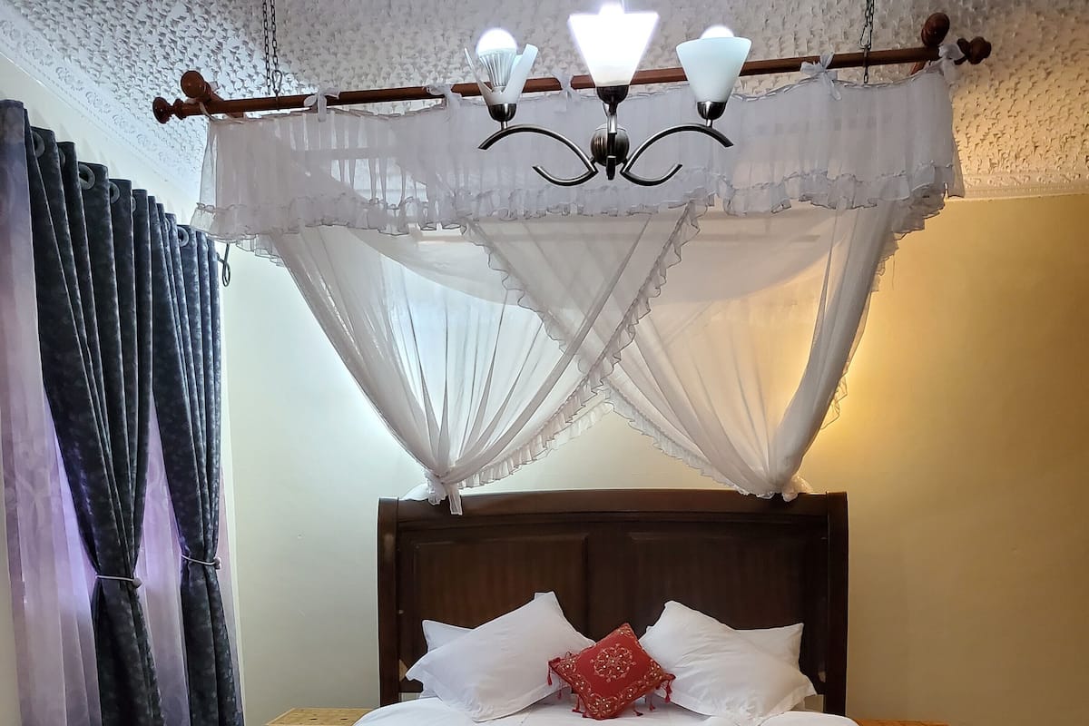 Una cama bien tendida con sábanas blancas y una almohada decorativa roja, un cabecero de madera y un dosel con una lámpara colgante.