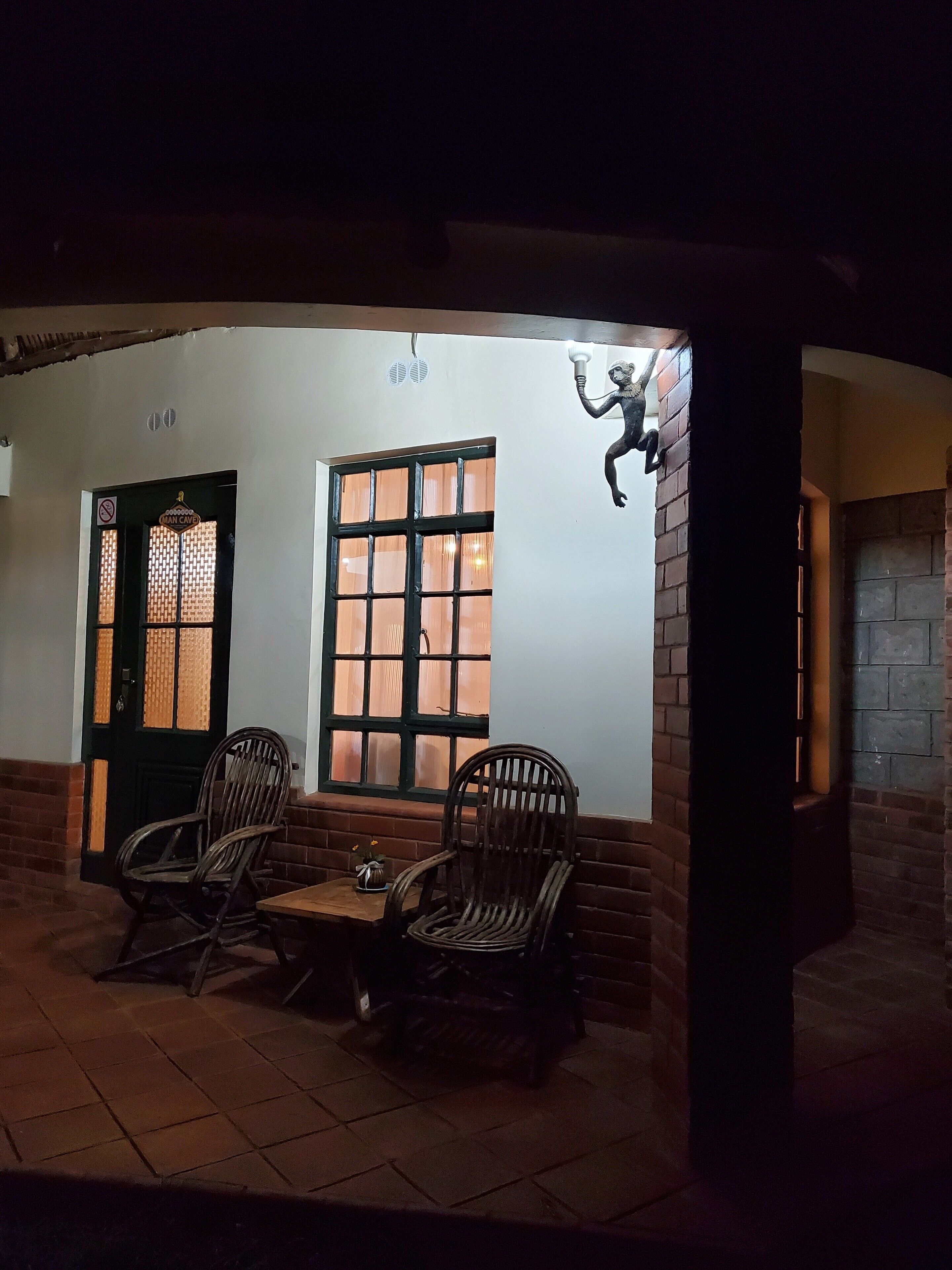 A covered outdoor seating area with two wicker chairs and a small table, a door with a 'No Entry' sign, and a wall-mounted sculpture.
