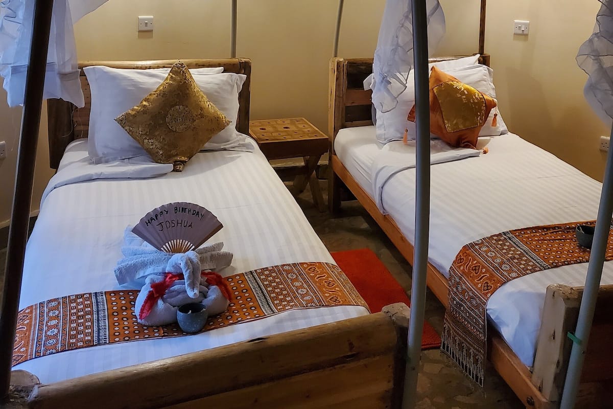 Una habitación con dos camas de madera, cada una con cubrecama blanco y manta estampada. Hay almohadas en las camas y una mesita de noche con una lámpara entre ellas.