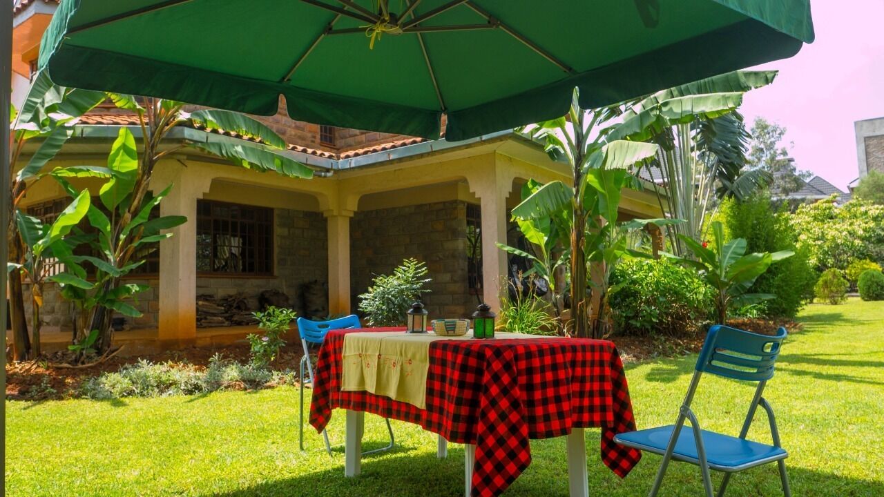 Een terras buiten met een tafel, afgedekt met een rood-zwart geruit tafelkleed, een groene parasol en een blauwe stoel.