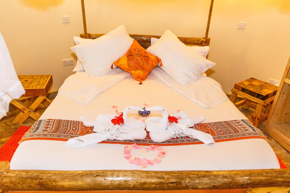 Una cama bien tendida con sábanas blancas, un cojín naranja decorativo y un arreglo de toallas sobre una colcha estampada.