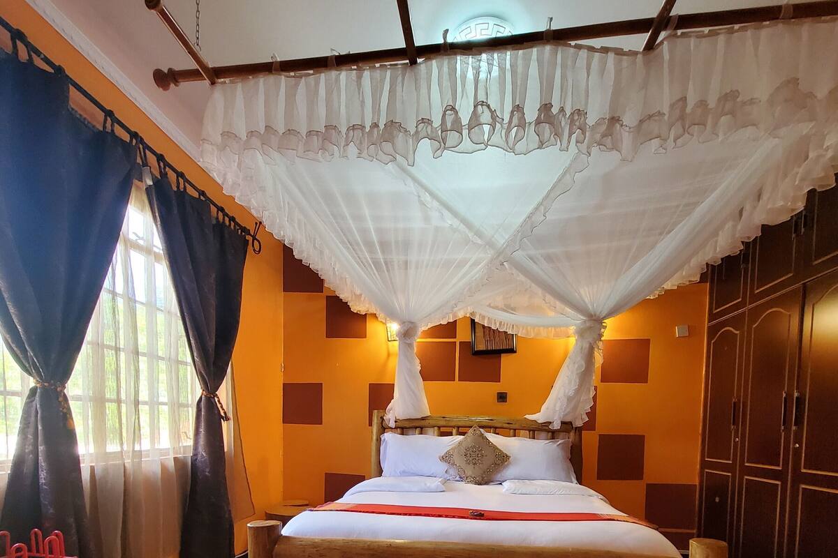 Una habitación con una cama bajo un dosel, un banco de madera, una ventana con cortinas y una pared con un diseño geométrico.