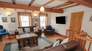 A living room with a brown leather sofa, a wooden coffee table, and a wooden staircase. There are two windows with curtains, a flat-screen TV on the wall, and a wooden door.