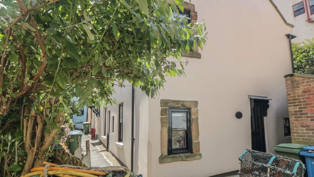 A narrow alleyway with a building on one side and a tree on the other. There is a window with a stone frame, a door, and a small balcony.