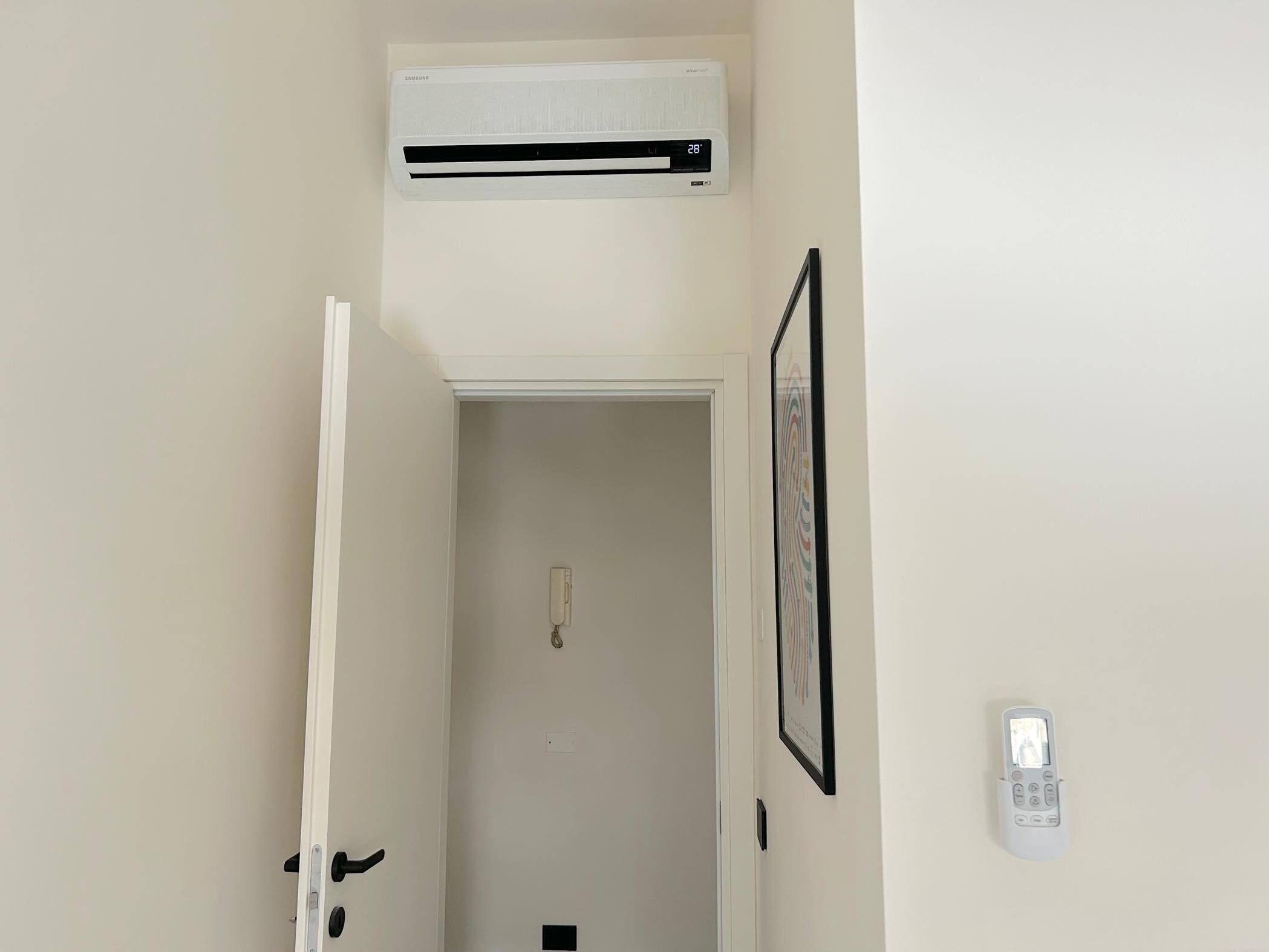 Een kamer met een witte deur, een wandairconditioner, een lijst met een afbeelding en een intercom bevestigd aan de muur.