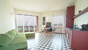 A modern apartment with a green sofa, a dining table with chairs, a kitchen area, and a balcony with a crane view.