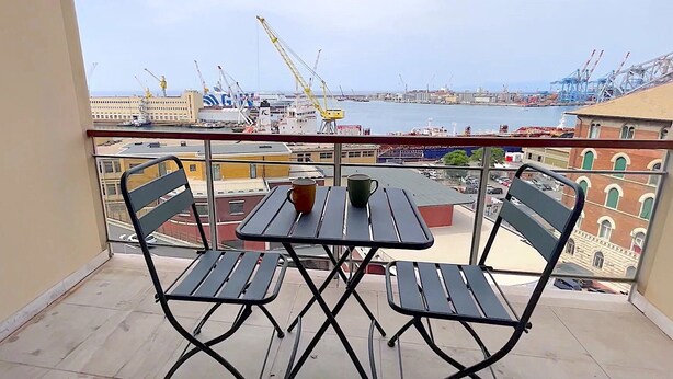 A balcony with two chairs and a table, overlooking a port with cranes and a large ship.