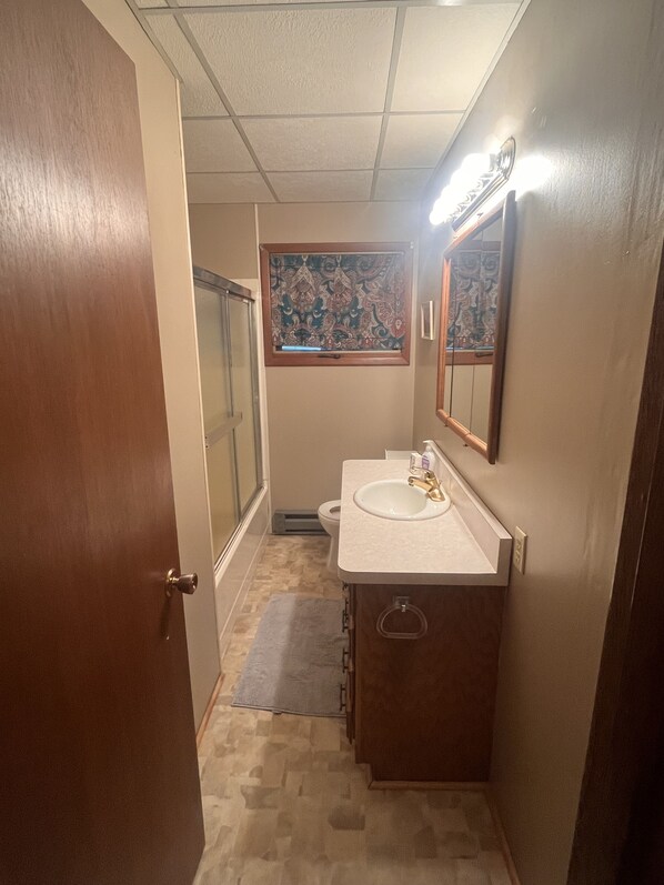 A bathroom with a wooden door, a sink with a mirror, a patterned window, and a shower area.