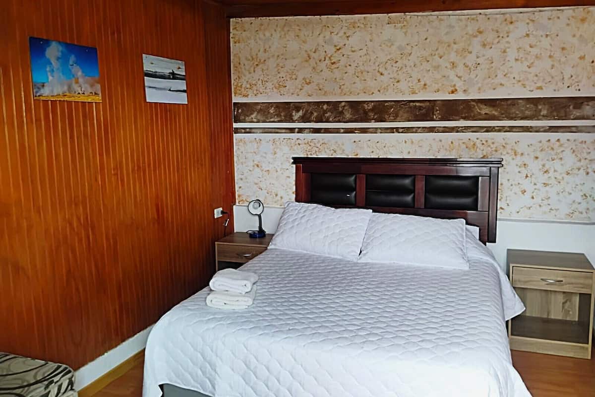 Quarto com parede de madeira, cama com roupa de cama branca, mesa de cabeceira com abajur e pequena mesa com espelho.