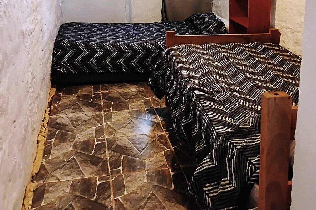 Quarto pequeno com duas camas, janela com cortinas, prateleira de madeira e piso com padrão de pedra.
