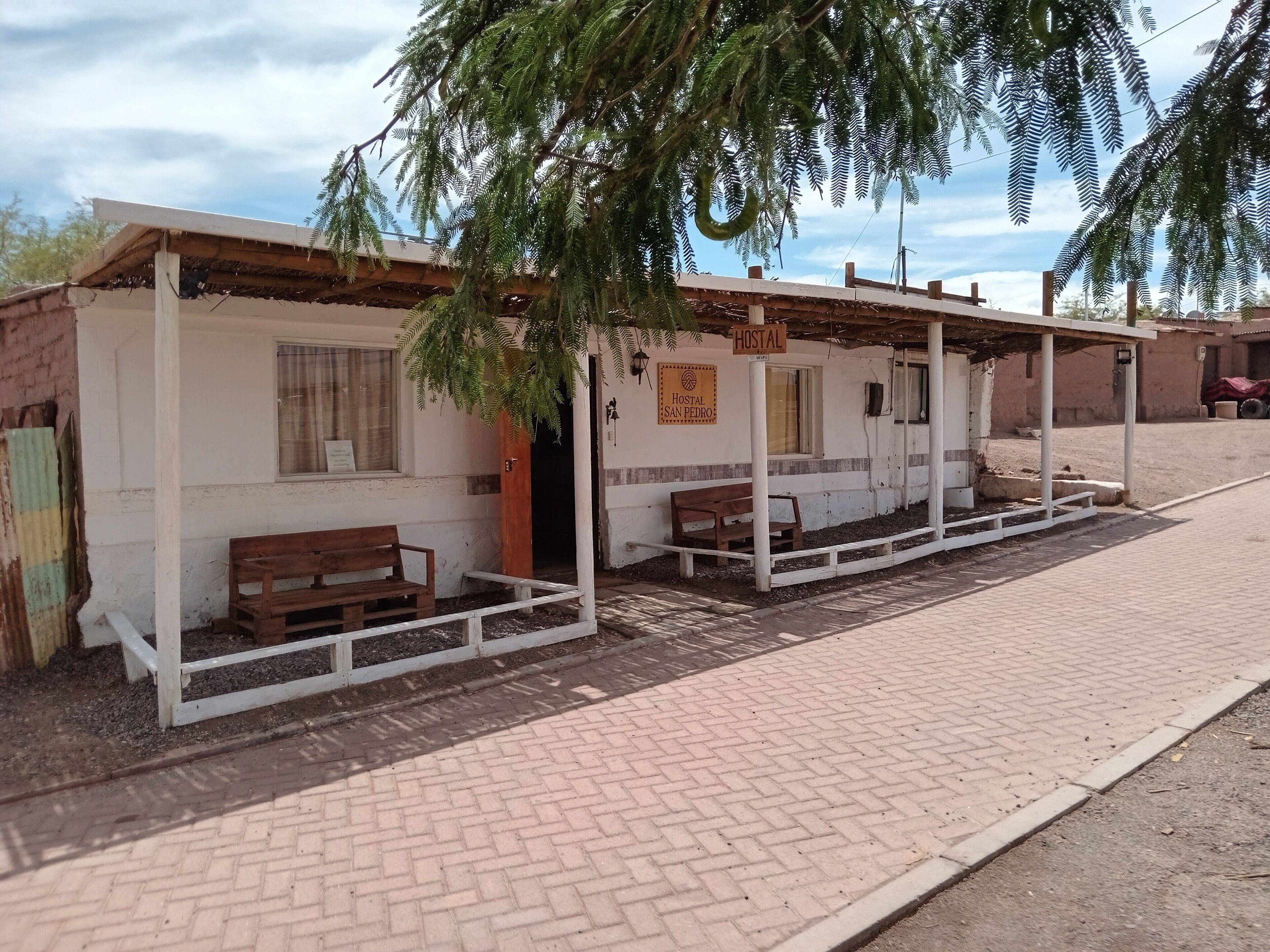A hostel with a sign reading "Hostal San Pedro," featuring a simple brick structure, wooden benches, and a covered seating area.