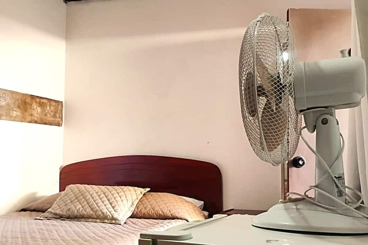 Quarto com cama, mesa de cabeceira, ventilador e vigamento de madeira no teto.