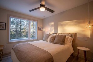 A bedroom with a large bed, a ceiling fan, a window with a view of trees, and a small table. - Panoramic view - Spa - Chalet du Cap (Matawinie Regional County Municipality)