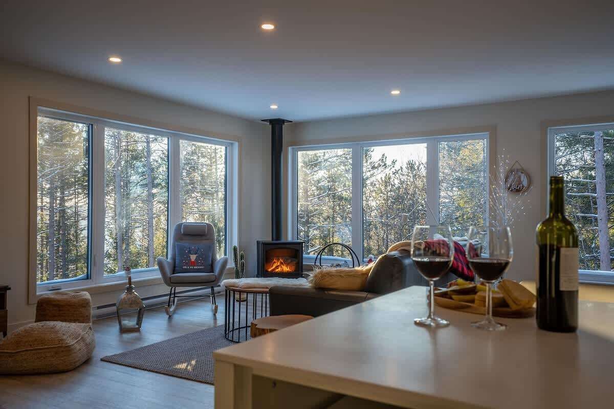 Ein modernes Wohnzimmer mit großen Fenstern, einem Kamin, bequemen Sitzgelegenheiten und einem Essbereich mit Weingläsern.