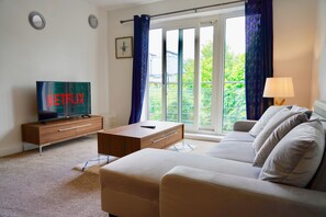 Relax in comfort with a flat-screen TV, sofa, and floor lamp, surrounded by a peaceful view of greenery.