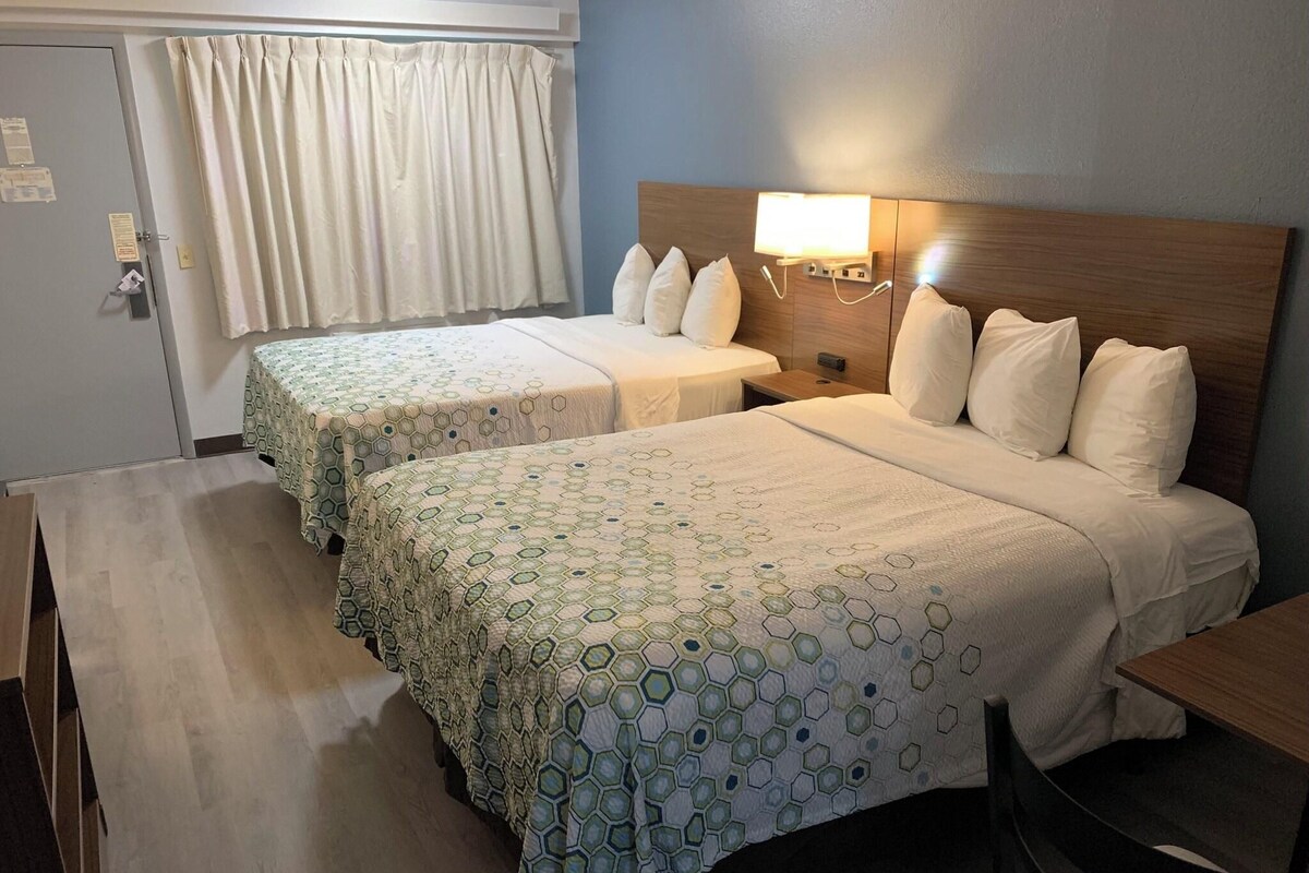 Een hotelkamer met twee bedden, een bureau, een stoel, een nachtkastje, een lamp en een raam met gordijnen.