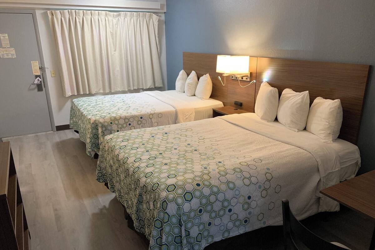 Een hotelkamer met twee bedden, een bureau, een stoel, een nachtkastje, een lamp en een raam met gordijnen.