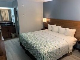 Habitación de hotel con cama, mesita de noche, lámpara y baño con lavabo y espejo.