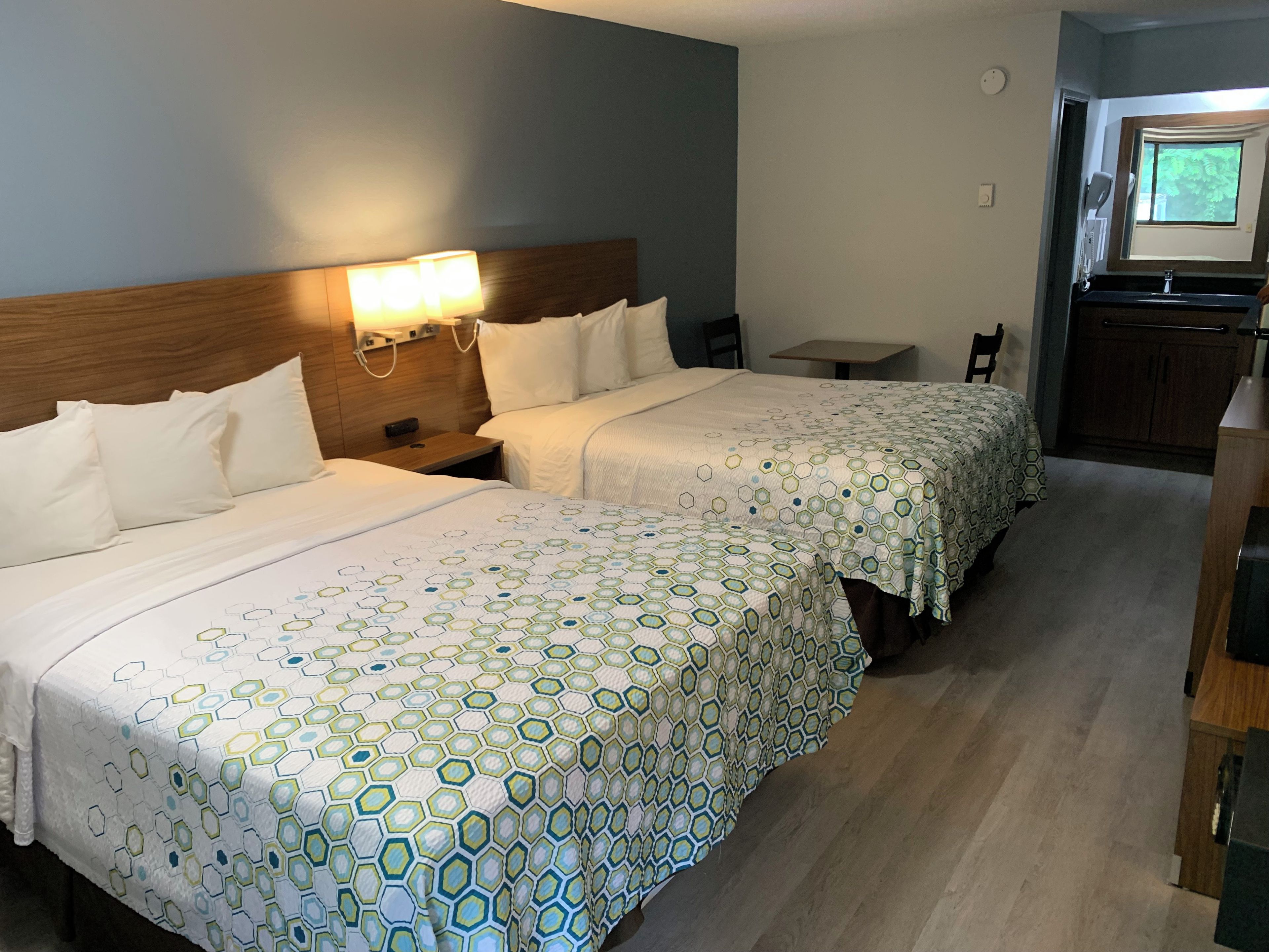 A hotel room with two beds, a small table, a chair, and a kitchenette area.