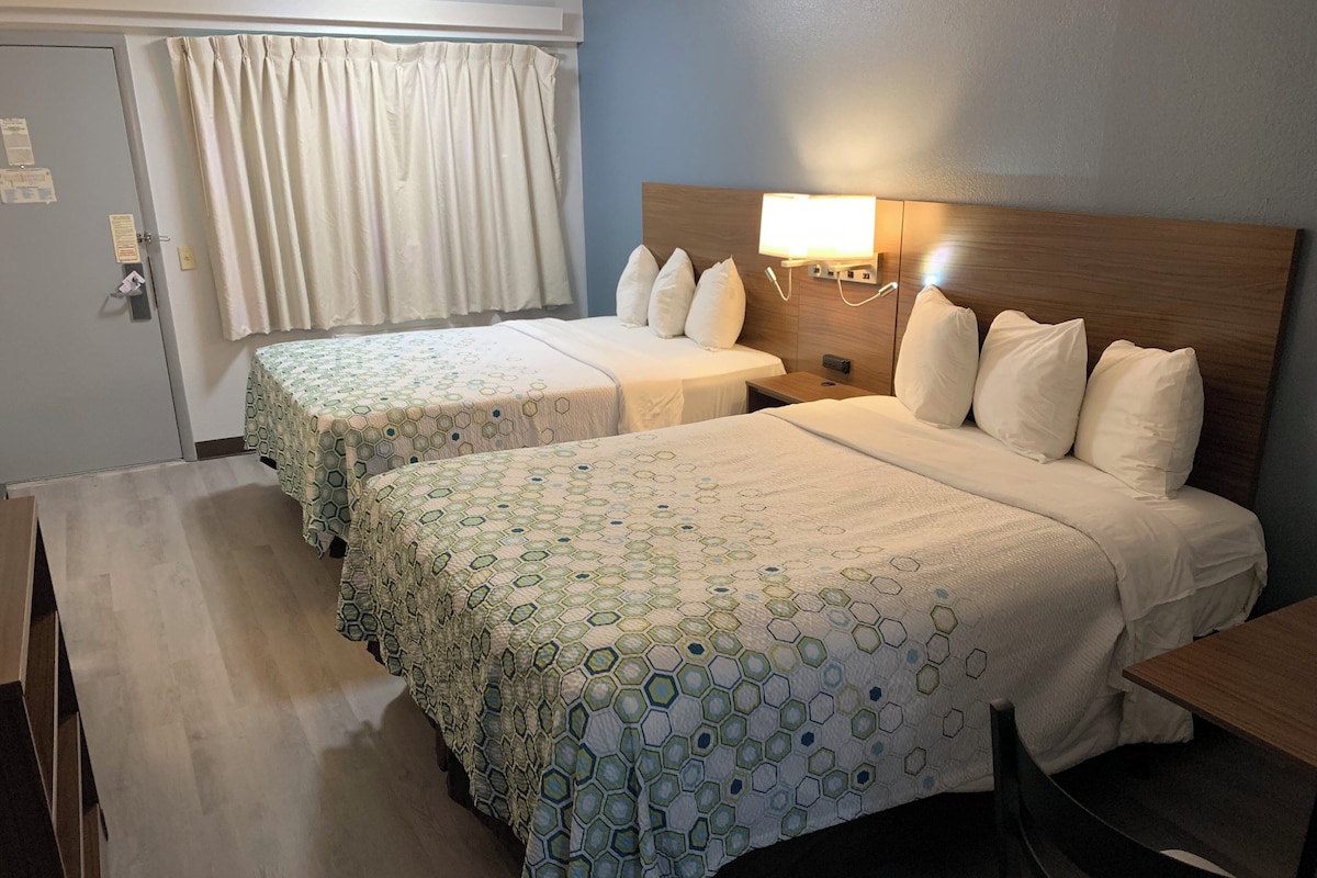 A hotel room with two beds, each with a hexagonal patterned bedspread, a wooden headboard, a bedside lamp, and a small table.