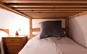 A small, single-bed room with a wooden bunk bed structure, a bedside table with a lamp, and a nightstand with a drawer.