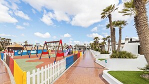Un parque infantil con columpios y toboganes, rodeado de palmeras y una cerca blanca.