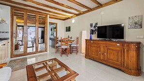Amplia sala de estar con techo de madera, comedor con mesa redonda y sillas, televisor sobre un mueble de madera y puerta corrediza de vidrio que da a un espacio exterior.