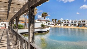 Una terraza de madera con vistas a un cuerpo de agua tranquilo, con una fila de edificios blancos al fondo.