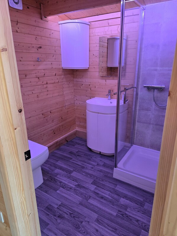 A small, compact bathroom with wooden walls, a white sink, and a glass-enclosed shower. - Pet Friendly Private Cabin Town Eurotunnel Beach (Hythe)