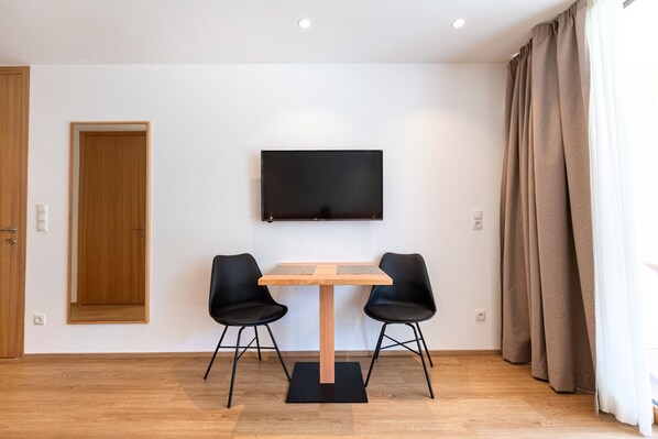 Une pièce moderne avec une table en bois et deux chaises noires, un téléviseur à écran plat fixé au mur et une porte en bois.