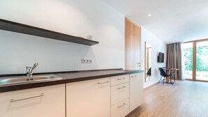 A modern kitchen with white cabinets, a black countertop, and a wooden shelf. There is a dining area with a table and chairs visible through a glass door. - 192A Greva Apartments 2 (Santa Cristina Valgardena)