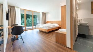 Una habitación de hotel moderna con una cama de madera, un escritorio con una silla, un ventanal con cortinas y un baño con lavamanos y espejo.