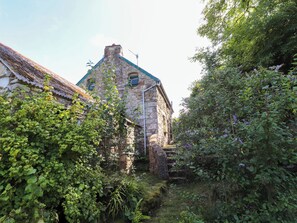 A stone building with a sloped roof, surrounded by dense greenery and a small stream. - Penfeidr Newydd (Newport)