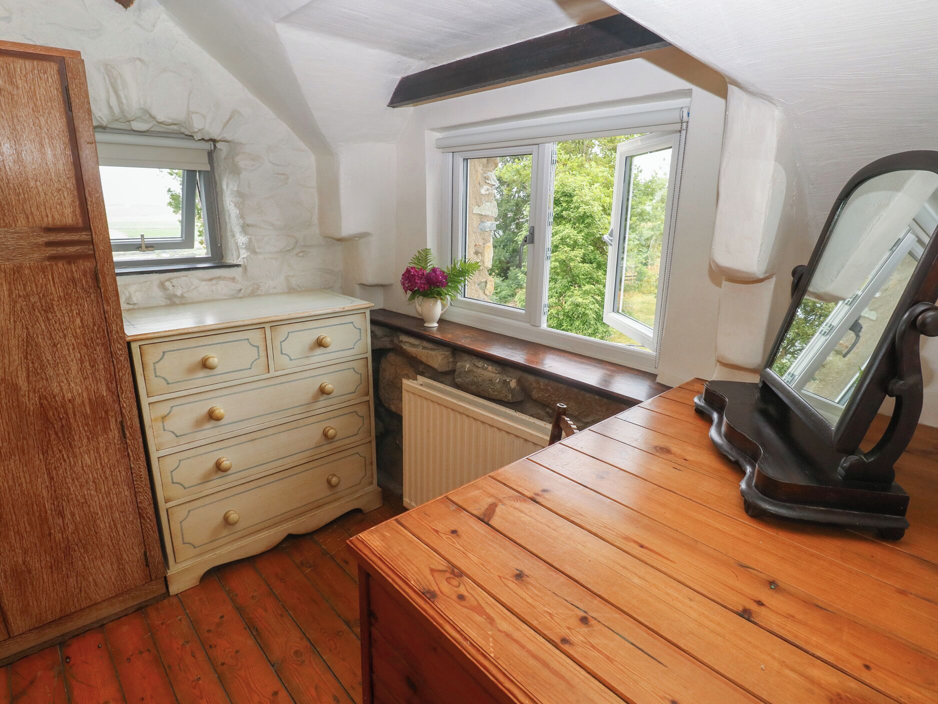 Una habitación con tocador de madera, un espejo, una ventana con vista a zonas verdes y un radiador.