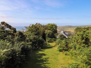 A grassy pathway leading through dense greenery towards a stone building with a chimney, overlooking a coastal landscape. - Penfeidr Newydd (Newport)