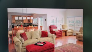 Una sala de estar con un sofá color beige, un reposapiés rojo y una mesa de centro. Se observa una cocina al fondo con armarios de madera y una zona de comedor con sillas.