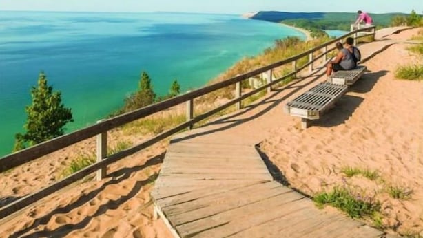 En træpromenade fører hen til et sandområde med udsigt over et stort vandområde og fjerntliggende land.